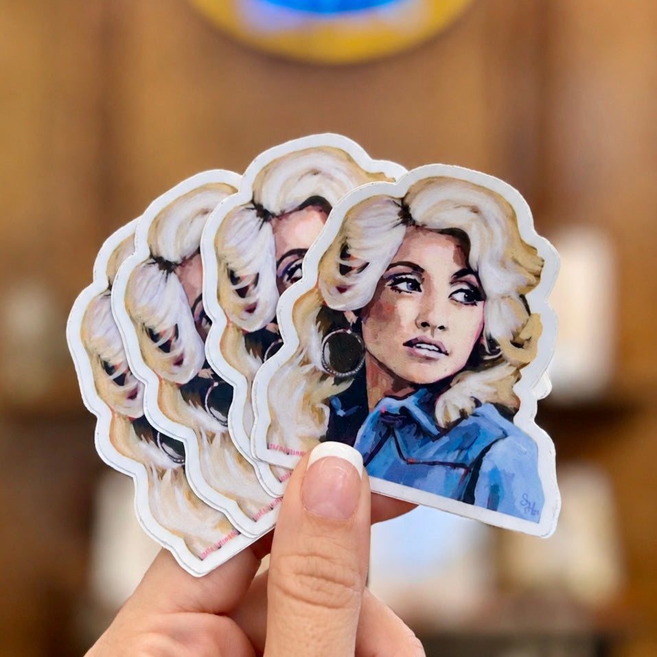 A hand holds a fan of die-cut stickers featuring a stylized portrait of a woman with blonde hair and a blue top, against a blurred warm-toned indoor background.