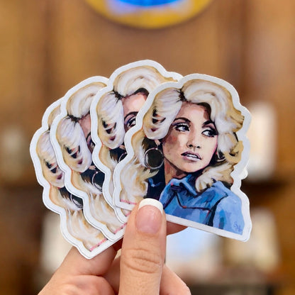 A hand holds a fan of die-cut stickers featuring a stylized portrait of a woman with blonde hair and a blue top, against a blurred warm-toned indoor background.