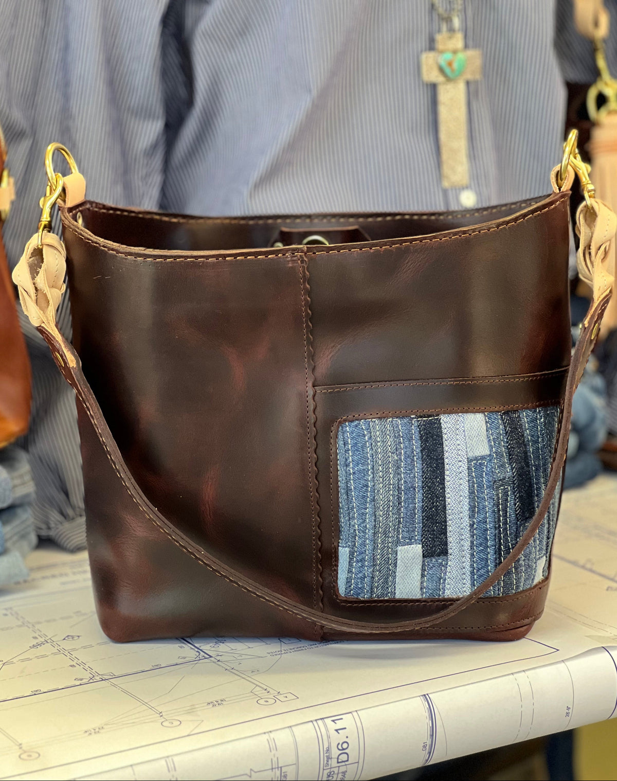 A dark brown leather shoulder bag with a blue and gray striped patchwork fabric pocket on the front, displayed on a table with a blue striped shirt in the background.